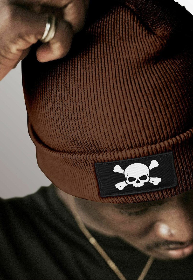 Brown ribbed beanie with a black patch featuring a white skull and crossbones design. Soft texture and a snug fit.