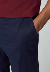 Bordeaux polo-shirt boven een navy broek. De broek heeft een gladde textuur, zijzak en gestikte naden. Een hand is zichtbaar in de broekzak.