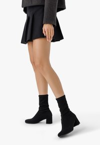 Botines negros con una parte superior de tejido suave y elástico, punteras cuadradas y tacones gruesos esculpidos, combinados con una falda negra plisada.