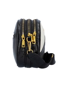 Sort og hvid crossover taske med tre guld lynlås og en sort justerbar stofrem, vist fra siden på en hvid baggrund.