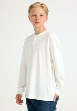 Weißes Langarm-Henley-Shirt mit Rundhalsausschnitt und Drei-Knopf-Leiste. Haptisch weicher Stoff und gerippter Stoff, kombiniert mit dunkelblauen Jeans.