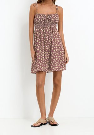 Woman wearing a sleeveless, knee-length purple dress with yellow floral pattern and tie straps, paired with beige flat sandals.