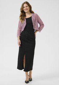 Gilet en dentelle rose clair sur un haut noir, associé à une jupe en denim noire avec une fente à l'avant et des talons noirs avec des accents.