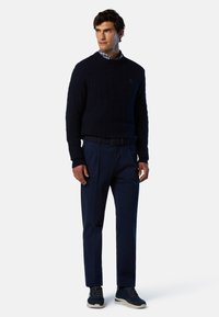 Maglione lavorato a maglia navy con design a treccia, abbinato a pantaloni navy scuri e sneakers navy. Il colletto della camicia a righe è visibile sotto il maglione.