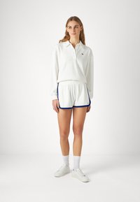 Bela polo majica z dolgimi rokavi in ovratnikom ter Lacoste logotipom, v kombinaciji z belimi kratkimi hlačami z modrim obrobjem. Maneken nosi bele superge in nogavice.