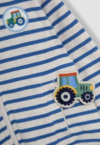Gestreifter Stoff in Creme und Blau, mit einem gestickten Traktor-Patch in Grün, Rot und Blau. Reißverschluss vorne.