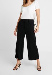 Zwarte wijde broek met een rechte pasvorm, hoge taille en frontzakken, gecombineerd met een beige top met korte mouwen en zwarte haksandalen.