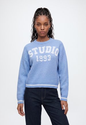 Jeune femme aux cheveux tressés portant un pull bleu avec un texte blanc "STUDIO 1993" et un jean foncé, debout devant un fond uni.
