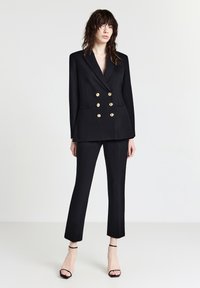 Blazer nero doppiopetto con bottoni dorati, abbinato a pantaloni neri su misura. Tacchi neri a strappo completano il look. Texture liscia.