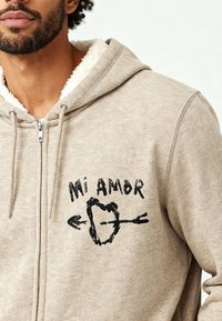 Sweat à capuche zippé beige avec doublure blanche douce, comportant le texte noir brodé "Mi Amor" et un design de cœur avec une flèche sur la poitrine.