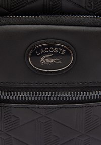 Sort Lacoste rygsæk med tekstureret geometrisk mønster, der har en sølvlogo-plade og en lynlåslukning med metallynlåstænder.