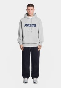 Szara bluza z kapturem z napisem "PROSTO." w kolorze granatowym, z sznurkiem i luźnym krojem, w zestawieniu z ciemnymi dżinsami i trampkami. Gładkie tło.