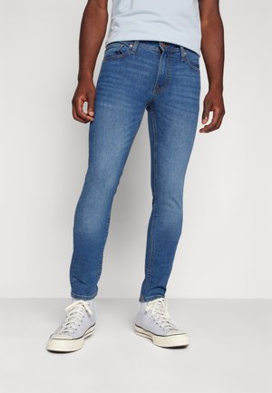 Mand iført slim-fit blå jeans og hvide høje sneakers, stående mod en ensfarvet lys baggrund.