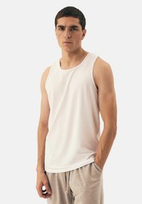 White sleeveless tank top with a round neckline, made of smooth fabric. Model wears light gray sweatpants, hands in pockets, neutral background.