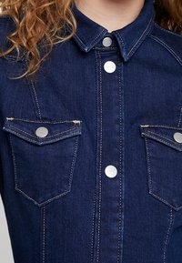 Tummansininen farkkutakki, jossa on nappikiinnitys, kaksi rintataskua, terävä kaulus ja kontrastivalkoiset ompeluyksityiskohdat. Näkyvät hopeiset napit.