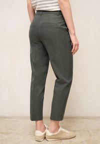 Kvinde iført mørkegrønne højtaljede skrå bukser og beige sneakers, stående mod en ensfarvet lys væg.