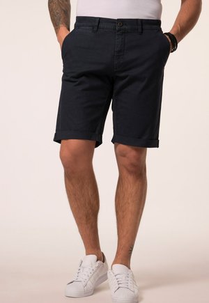 Mand iført sorte, op-rullede shorts, hvide sneakers og en hvid t-shirt, med hænderne i lommerne og synlige tatoveringer på arme og ben.