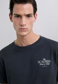Camiseta de algodón gris oscuro con cuello redondo y mangas cortas. Presenta un logotipo en texto blanco en la parte izquierda del pecho. Textura suave.