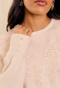 Gaiši rozā džemperis ar sarežģītu ziedu mežģīņu detaļu, ribotām piedurknēm ar trim dekoratīvām pogām un mīkstu tekstūru.