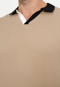 Gros plan d'une personne portant une chemise beige texturée avec un col noir et une patte intérieure blanche visible au niveau de l'encolure.