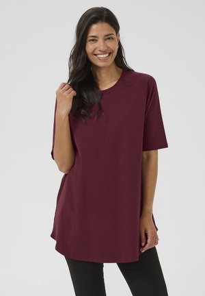 Bordeauxfarbene übergroße T-Shirt mit kurzen Ärmeln, Rundhalsausschnitt und seitlichem Schlitzsaum, getragen mit schwarzen slim-fit Hosen. Glatte Stofftextur.