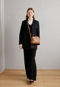 Sac en cuir marron au design structuré, avec un rabat à l'avant et des accessoires en argent, porté en bandoulière contre un costume rayé noir.