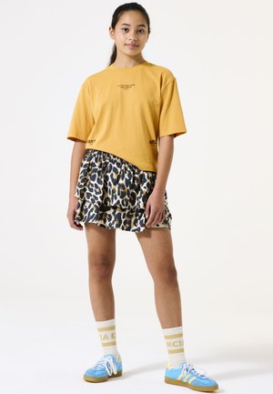 Geel cropped t-shirt met afgedrukte tekst, gecombineerd met een gelaagde leopardprint rok. Blauwe sneakers en gestreepte gele sokken maken de look compleet.