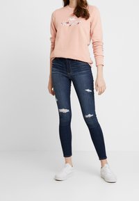 Ljusrosa sweatshirt med blommönsterbroderi, ihopparad med mörkblå skinny jeans med slitna revor, kompletterad med vita sneakers.