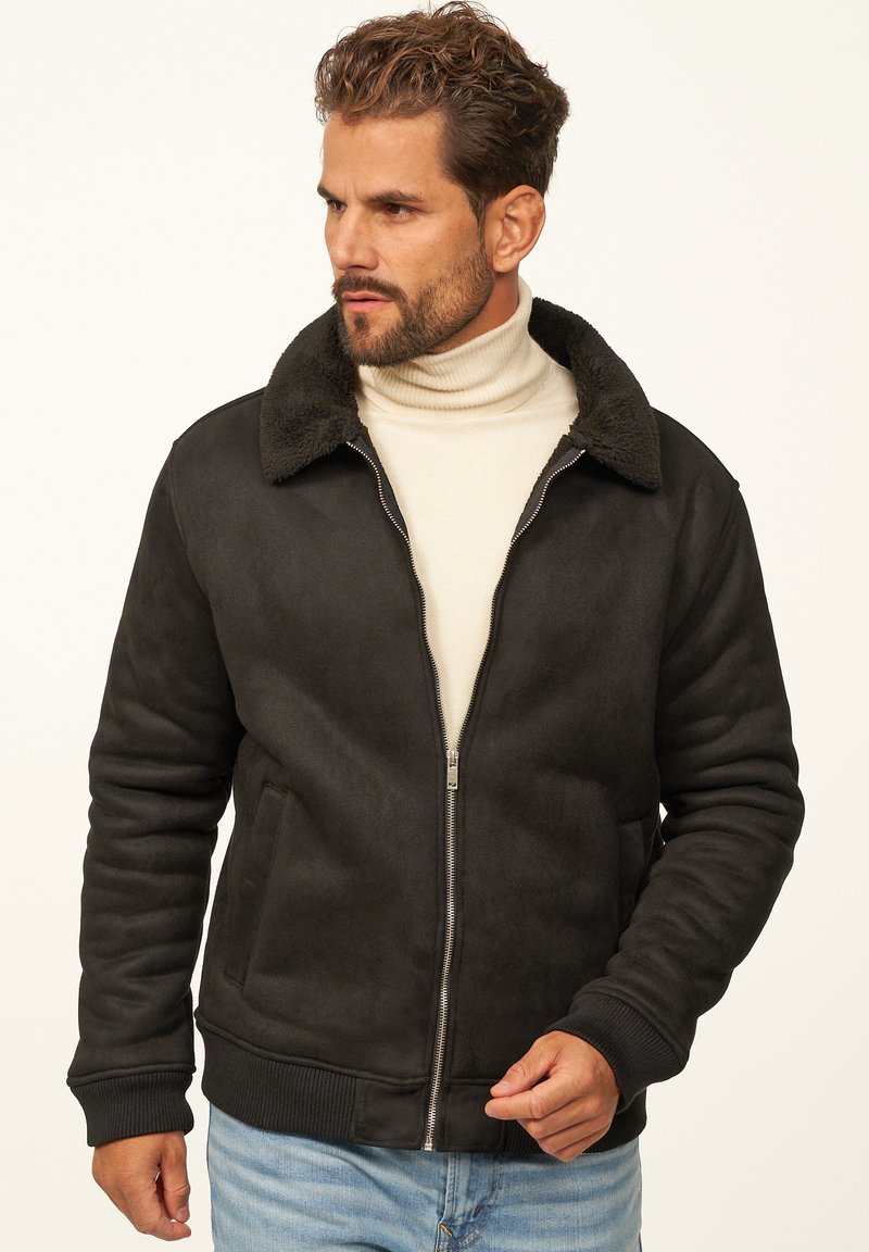 Schwarze Wildleder-Bomberjacke mit einem strukturierten, Kunstfellkragen. Verfügt über einen Reißverschluss vorne, gerippte Bündchen und seitliche Taschen. Wird über einem cremefarbenen Rollkragenpullover getragen.