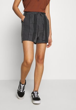Shorts a righe bianche e nere con cintura annodata e tasche laterali, abbinati a scarpe da ginnastica nere con dettagli bianchi.