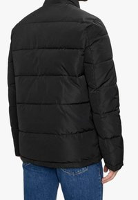 Persona vestida con una chaqueta acolchada negra y jeans azules, vista desde atrás contra un fondo blanco sencillo.