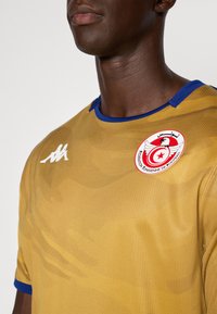 Kuldne spordijerseysin siniste aktsentidega, millel on tekstureeritud kangas ja ümmargune kaelus. Ülemises vasakus rinnakus on ümar logo.