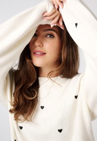 Crème trui met zwarte hartpatronen, met lange, losse mouwen. Het model heeft golvend bruin haar en draagt een eenvoudige zilveren ketting.