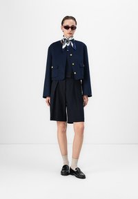 Persoană îmbrăcată cu o jachetă scurtă bleumarin cu nasturi aurii, pantaloni scurți negri, eșarfă cu model la gât, șosete bej, mocasini negri și ochelari de soare roșii.