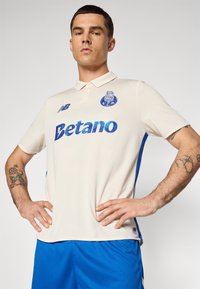 Polo créme avec des accents bleus, arborant un logo sur la poitrine et le mot "Betano" en texte bleu irisé. Manches courtes et tissu texturé.