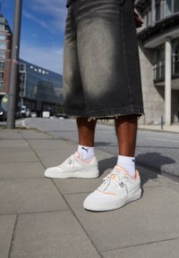 Białe sneakersy z pomarańczowymi akcentami, teksturowana tkanina i gładka gumowa podeszwa. Noszone z czarnymi szortami dżinsowymi do kolan z postrzępionymi brzegami i białymi skarpetkami.