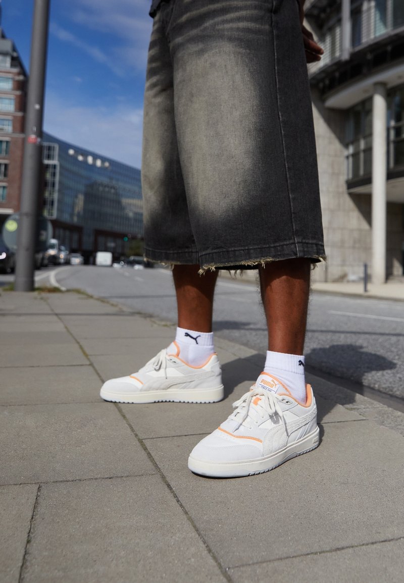 Białe sneakersy z pomarańczowymi akcentami, teksturowana tkanina i gładka gumowa podeszwa. Noszone z czarnymi szortami dżinsowymi do kolan z postrzępionymi brzegami i białymi skarpetkami.