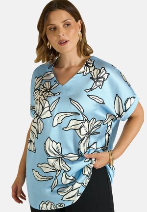 Kvinde iført lyseblå bluse med stort hvidt og sort blomsterprint og sorte bukser, accessoriseret med guldringe og armbånd.