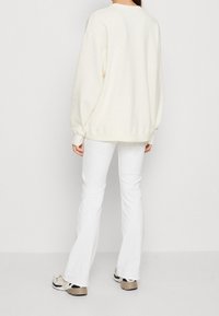 Krémová mikina s volným střihem, kulatým výstřihem a ribkovanými manžetami. V kombinaci s bílými zvonovými kalhotami a vzorovanými teniskami. Zadní pohled.