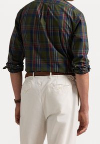 Camicia a quadri verdi con accenti blu e bordeaux, maniche arrotolate, abbinata a pantaloni crema con tasche posteriori. Cintura marrone visibile.