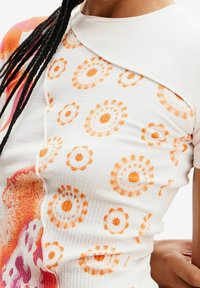 Fitted vit topp med ribbad textur som har orange blommönster och rund krage. Designen inkluderar flerfärgade detaljer på sidan.