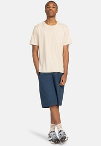 Béžové bavlnené tričko s prednou vreckom, spojené s námornícko modrými šortkami. Dokončené vzorovanými teniskami a svetlými ponožkami.