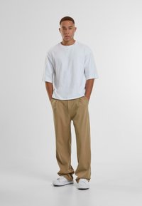 Hvit oversized t-skjorte, beige plissert bukse og hvite sneakers. Modellen står med hendene i lommene mot en ensfarget bakgrunn.