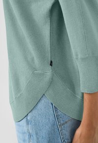 Zbliżenie na osobę w jasnozielonym swetrze z małą czarną metką, noszącą jasnoniebieskie dżinsy.