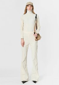 Mono beige ajustado con cuello alto, costuras contorneadas, combinado con una gorra beige y un pequeño bolso marrón. Zapatos negros de punta completan el conjunto.