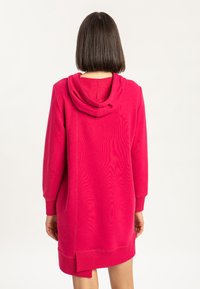 Kobieta z prostymi ciemnymi włosami ma na sobie jasnoróżowy długi sweatshirt z kapturem, pokazana od tyłu na jednolitym białym tle.