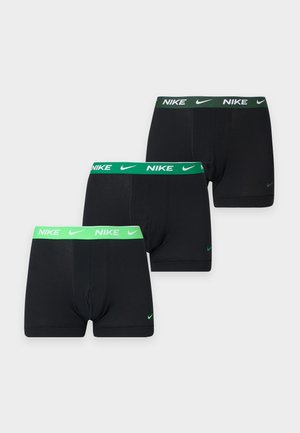 Sale tumedama nahaga mees seisab paljajalu, kandes musti Nike bokserpükse tumerohelise vöökohaga, helel ühevärvilisel taustal.