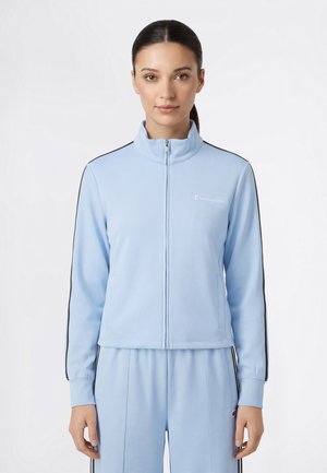 Donna che indossa una giacca sportiva azzurra con zip e pantaloni abbinati con strisce nere e logo Champion, in piedi davanti a uno sfondo bianco.