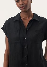 Camicia nera a maniche corte con bottoni, realizzata in tessuto testurizzato, con colletto, due tasche sul petto e bottoni neri.