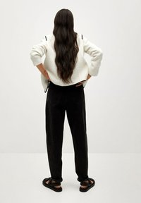 Nainen, jolla on pitkät tummat hiukset, seisoo kädet lanteilla valkoisessa tekstuurilla varustetussa takissa, mustissa housuissa ja mustissa sandaaleissa yksinkertaista taustaa vasten.